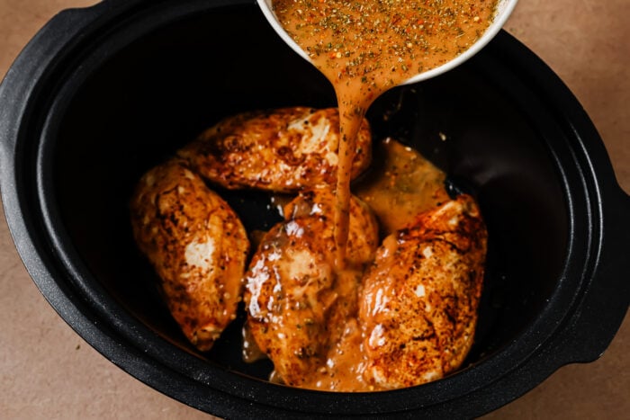Herbed sauce being poured from a bowl over seasoned chicken breasts in a slow cooker.