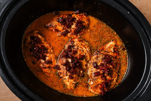 Three chicken breasts cooking in a slow cooker, topped with sun-dried tomatoes and herbs in a flavorful broth.