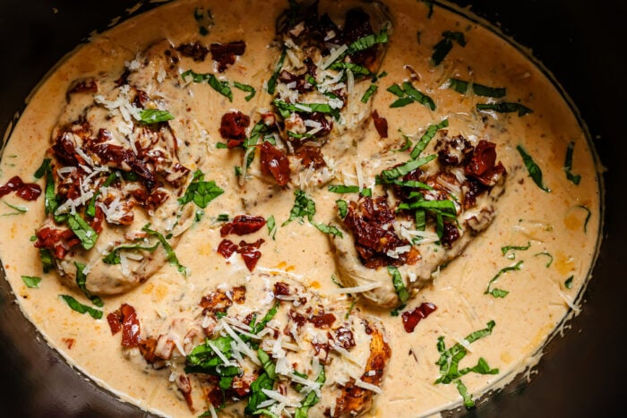 Chicken breasts simmering in a creamy sauce with sun-dried tomatoes, shredded cheese, and fresh basil.