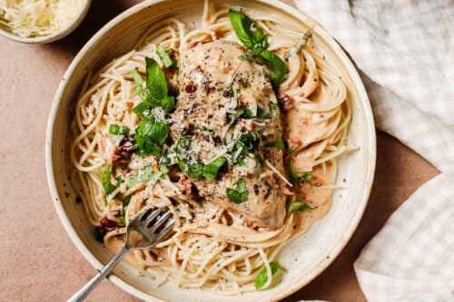 A bowl of creamy Crock Pot Marry Me Chicken pasta topped with a chicken breast, fresh basil, and shaved cheese, with a fork resting in the noodles.