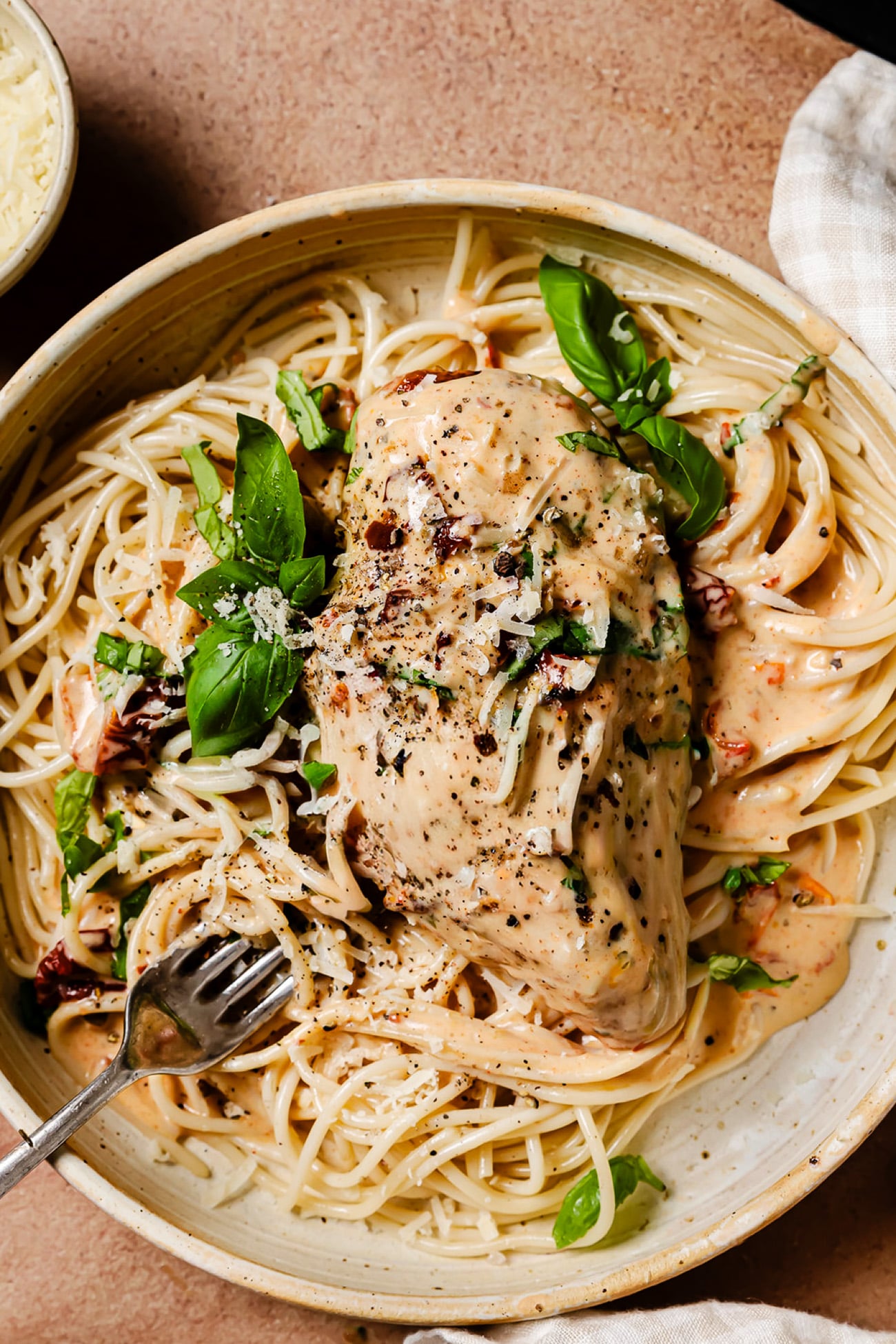 A bowl of creamy pasta topped with a chicken breast, sun-dried tomatoes, and fresh basil.