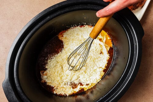 A hand uses a whisk to mix shredded cheese into a sauce inside a slow cooker.