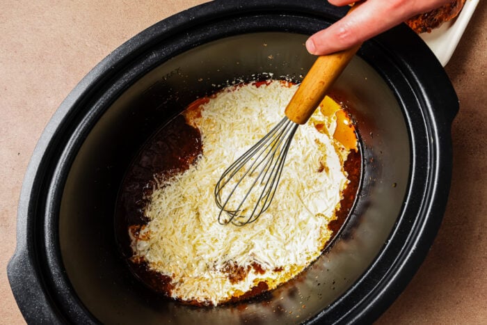 A hand uses a whisk to mix shredded cheese into a sauce inside a slow cooker.