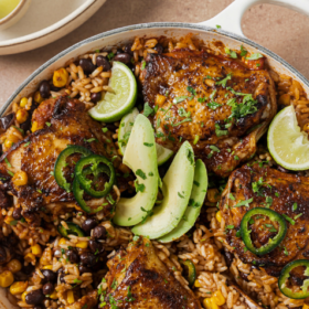 Skillet of Mexican chicken, rice, black beans, and corn, garnished with avocado, lime wedges, and jalapeño slices.