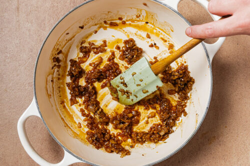 A hand uses a silicone spatula to stir browned, sautéed aromatics in a white enameled Dutch oven.