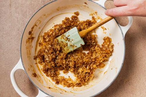 A hand uses a spatula to stir rice coated in a dark sauce in a white enameled pot.