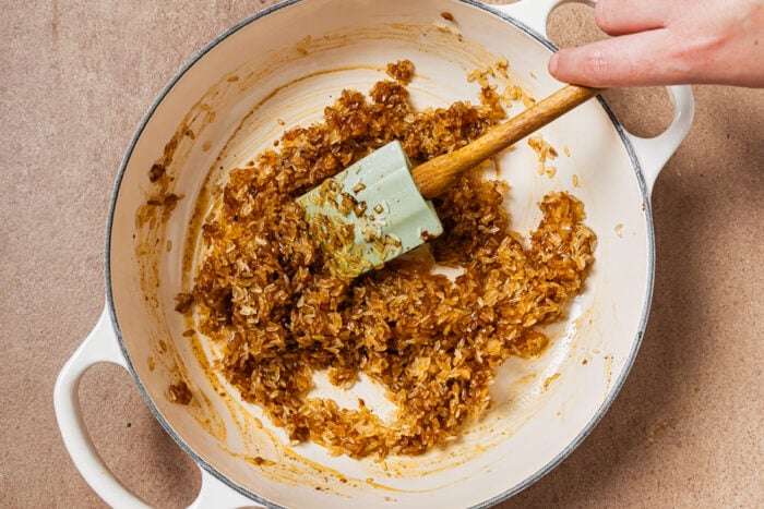 A hand uses a spatula to stir rice coated in a dark sauce in a white enameled pot.