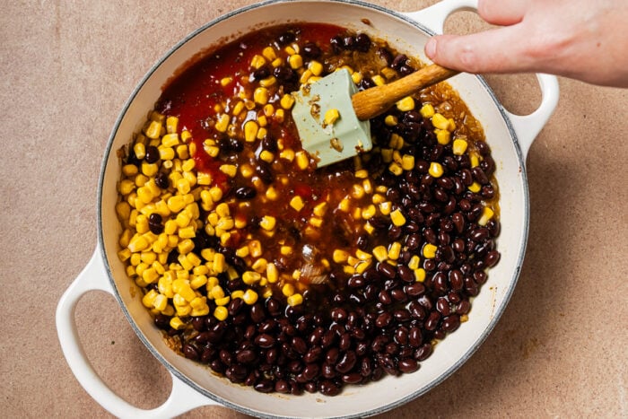 A hand uses a spatula to stir black beans, corn, and red sauce in a wide white pan.