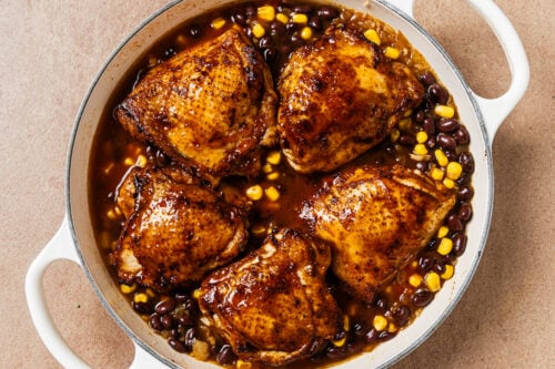 Four seared chicken thighs served in a skillet with black beans and corn in a dark sauce.