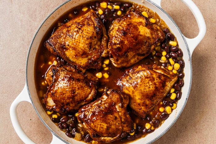 Five seared chicken thighs served in a skillet with black beans and corn in a dark sauce.
