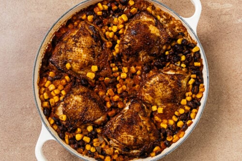 A white skillet filled with four seared chicken thighs cooked with black beans and corn.