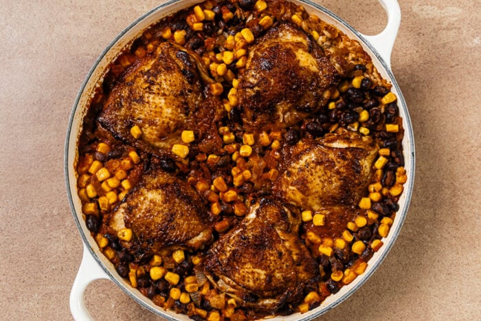 A white skillet filled with five seared chicken thighs cooked with black beans and corn.
