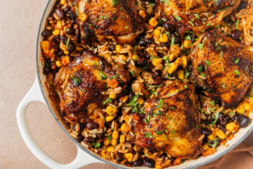 A skillet filled with seared chicken thighs, black beans, corn, and rice, garnished with fresh herbs.