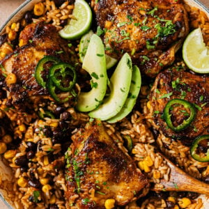 Skillet meal of glazed chicken thighs served over rice, black beans, and corn, garnished with avocado slices, jalapeño rings, and lime wedges.