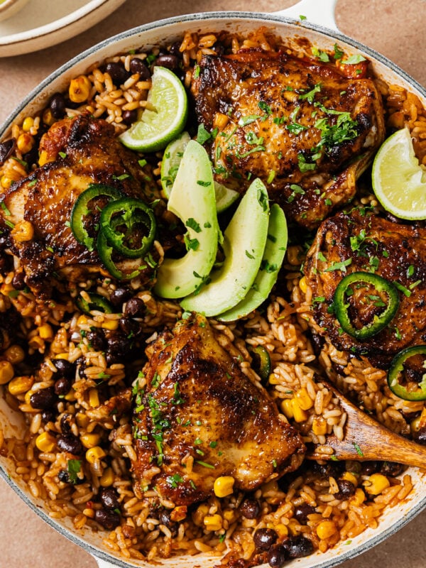 Skillet meal of glazed chicken thighs served over rice, black beans, and corn, garnished with avocado slices, jalapeño rings, and lime wedges.