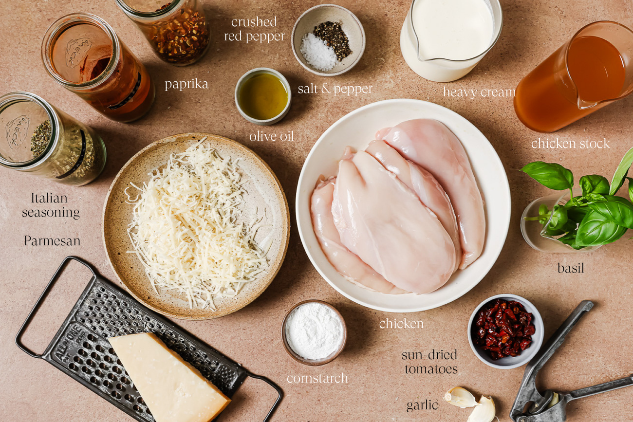 Ingredients for a Crock Pot Marry Me Chicken recipe laid out on a table, including chicken breasts, parmesan, heavy cream, chicken stock, sun-dried tomatoes, basil, garlic, cornstarch, olive oil, spices, and a grater.